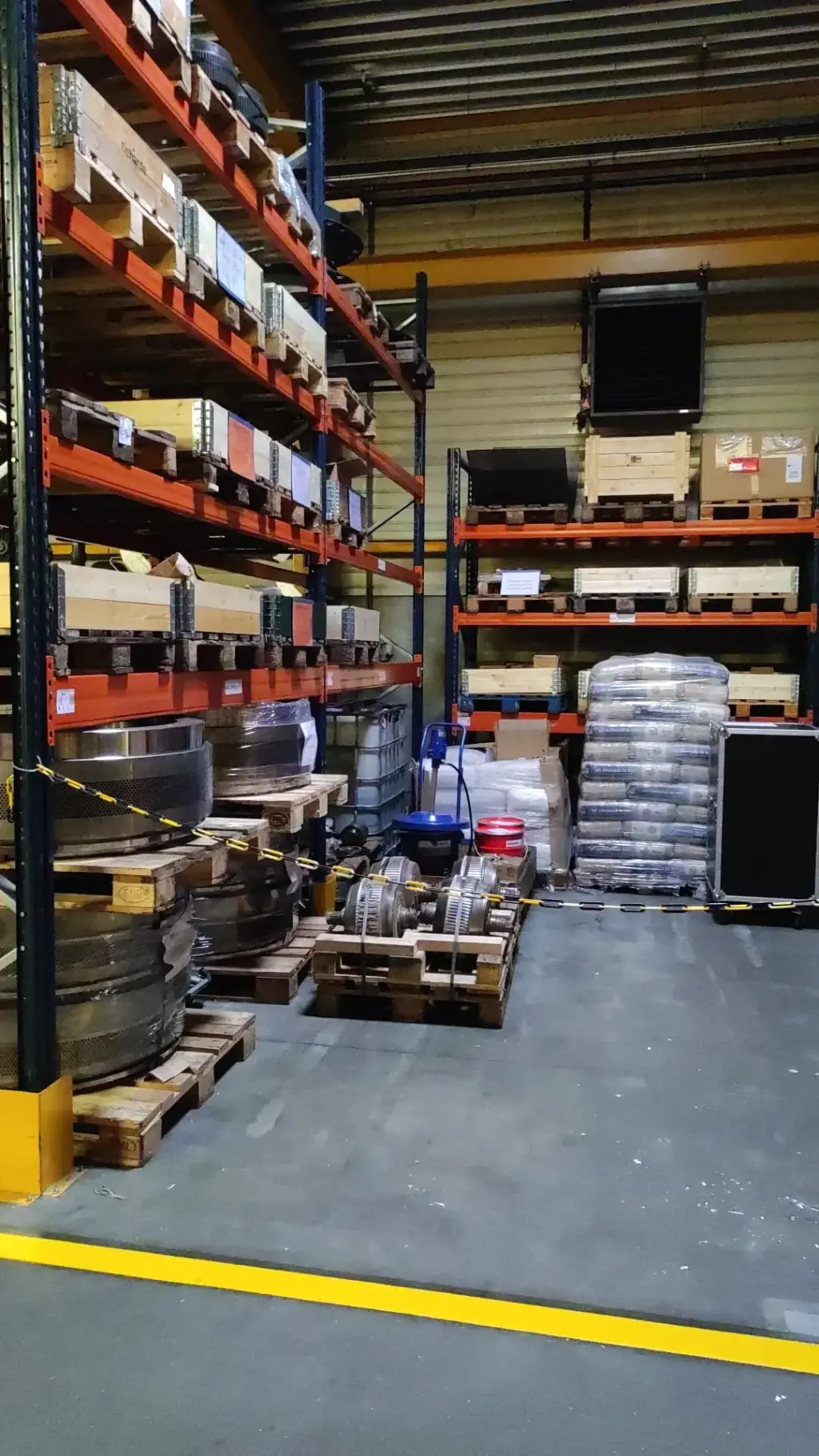 Industrielles Lagerhaus mit orangefarbenen Metallregalen, Kisten, Motoren und verpackten Materialien auf Paletten.